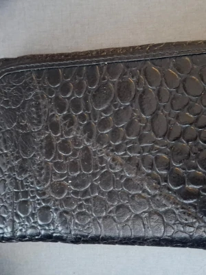 Svart plånbok med krokodilmönster - Cool svart plånbok med krokodilmönstrad yta och flera fack för kort och kontanter. Plånboken har dragkedja på insidan och är tillverkad i ett läderliknande material. Perfekt för dig som gillar stilrena accessoarer med lite edge.