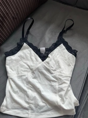 Vit linnetopp med spetsdetaljer - Snygg vit linnetopp med svarta spetsdetaljer längs urringningen och tunna axelband. Toppen har en omlott-look framtill och är tillverkad i mjukt material med stretch. Perfekt att styla med jeans eller kjol för en trendig look.