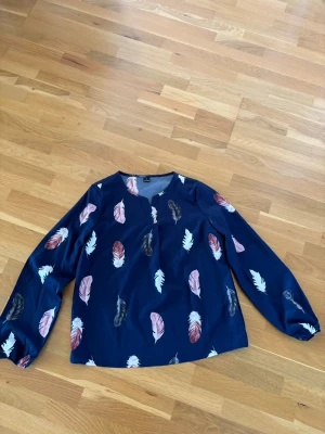 Blus med fjädermönster  - Långärmad blus med rund hals och liten öppning framtill. Mönstret med fjädrar i vitt, rosa och grått ger ett stilrent men samtidigt lekfullt uttryck. Resår vid är sluten för en snygg och bekväm passform. 