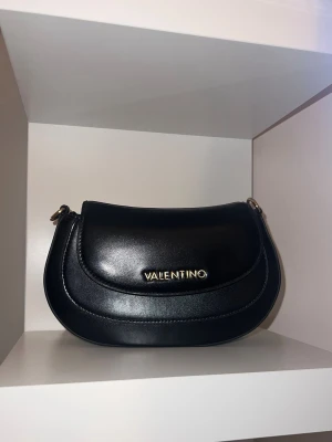  Valentino väska - Snygg svart handväska från Valentino med guldfärgad logga framtill. Väskan har rundad form och justerbar axelrem. Perfekt för dig som gillar stilrena accessoarer med exklusiv känsla. Pris kan diskuteras!!!!