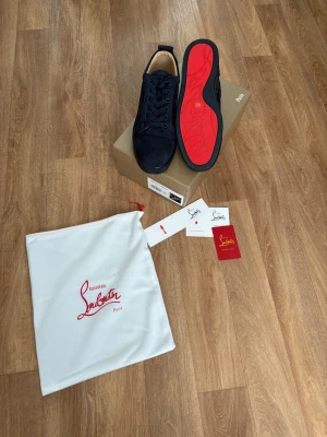 Mörkblåa sneakers från Christian Louboutin - Snygga svarta sneakers från Christian Louboutin med klassisk röd sula och diskret logga. Skorna har en låg profil, rund tå och är tillverkade i ett lyxigt material som känns exklusivt. Perfekt för dig som vill ha något stilrent och trendigt.