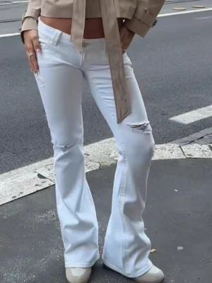 Vita bootcut jeans med slitningar - Snygga vita bootcut jeans med slitningar på låret och knät. Jeansen har hög midja och klassisk femficksdesign. Perfekta för en trendig och avslappnad look. Materialet ser ut att vara bomullsdenim med lite stretch.