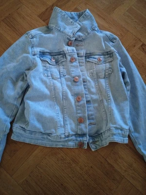 Ljusblå klassisk jeansjacka - Snygg ljusblå jeansjacka med klassisk krage och kopparfärgade knappar framtill. Två bröstfickor med lock och knapp, samt dekorativa sömmar. Perfekt för en avslappnad och trendig look.