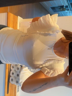 Vit topp med volangärmar - Säljer en söt vit topp med korta volangärmar och smockdetaljer vid axlarna. Toppen har rund halsringning och är figurnära i modellen. Perfekt för dig som gillar romantiska detaljer och vill ha en feminin look.