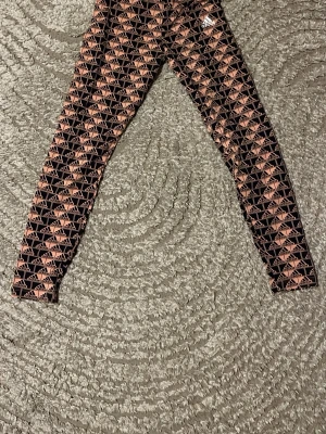 Svarta leggings med rosa Adidas-mönster - Säljer ett par svarta leggings från Adidas med all-over rosa logomönster. Tajt passform och elastisk midja, perfekta för en sportig och trendig look. Adidas-loggan syns tydligt framtill. Supersnygga till sneakers och hoodie.