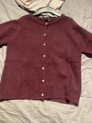 Vinröd kofta med pärlknappar 157 - Mysig vinröd kofta från 157 i storlek XS. Koftan har rund halsringning och stängs med snygga  pärlknappar framtill. Materialet är stickat och känns mjukt mot huden. Perfekt att slänga på sig när det är lite kyligt ute.