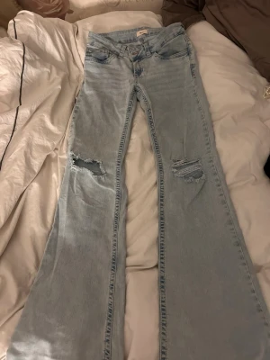 Ljusblå utsvängda jeans från Nelly - Säljer ett par ljusblå jeans från Nelly med utsvängda ben och slitna detaljer på båda låren. Jeansen har klassisk femficksdesign och dragkedja med knapp. Perfekta för dig som gillar en trendig och avslappnad look.