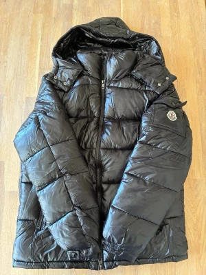 Moncler puffer jacka - Snygg svart pufferjacka från Moncler med glansig finish, huva och dragkedja framtill. Jackan har två framfickor med dragkedja och Moncler-logga på ärmen. Perfekt för kalla vinterdagar och riktigt trendig look.