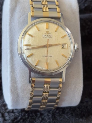 Vintage Tissot Seastar herrklocka – tvåton rostfritt stål med datum - Elegant Tissot Seastar herrklocka i klassisk vintage-stil. Tvåton rostfritt stålarmband med gulddetaljer och guldfärgad urtavla med datumfönster vid klockan tre. Mekanismen fungerar fint och klockan är i gott skick för sin ålder. Perfekt för dig som gillar tidlösa schweiziska klockor.