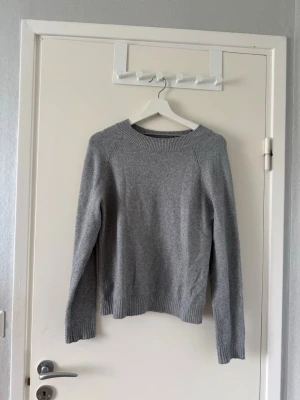 stickad tröja - En enkel och stilren ljusgrå stickad tröja med rund halsringning och långa ärmar. Tröjan har ribbstickade muddar vid ärmslut och nederkant. Perfekt att matcha med jeans eller kjol för en avslappnad look.