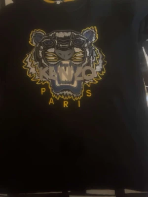 Svart Kenzo t-shirt med tigertryck - Säljer en svart t-shirt från Kenzo med stort broderat tigerhuvud i grått, blått och gult på bröstet. Texten 'KENZO PARIS' är broderad under motivet. Klassisk passform och rund halsringning. Perfekt statement-plagg för dig som gillar streetwear.