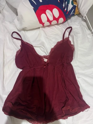 Vinrött baby doll topp med spets från Nelly - Mörkrött baby doll topp från Nelly med tunna axelband och fina spetsdetaljer längs urringning och fåll. Mjuk och lätt tygkänsla, liten rosett framtill. 