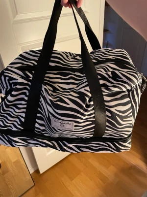 Zebramönstrad weekendbag Zuomanni - Snygg och rymlig weekendbag från Zuomanni med zebramönster i svart och vitt. Väskan har svarta handtag, dragkedja upptill och en praktisk ytterficka med dragkedja. Perfekt för resor eller gymmet, tillverkad i slitstarkt tygmaterial.