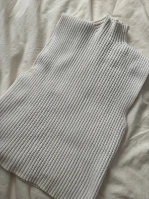 Vit ribbad ärmlös topp från Zara - Säljer en snygg vit ribbad topp från Zara med hög krage och ärmlös design. Perfekt att styla med jeans eller kjol för en clean look. Materialet är mjukt och stretchigt, vilket gör den riktigt bekväm att bära.