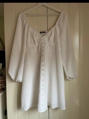 Vit klänning med knappar  - Säljer en vit kortklänning med v-ringad hals och långa ärmar. Klänningen har knappar framtill. Aldrig använd! Passar perfekt till student eller skolavslutning. 🌸