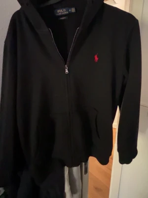 Svart hoodie från Polo Ralph Lauren - Snygg svart hoodie med dragkedja från Polo Ralph Lauren. Klassisk design med huva, två fickor framtill och det ikoniska röda Polo-logot på bröstet. Tillverkad i mjukt bomullsmaterial som är skönt att bära. Perfekt för en avslappnad och stilren look.