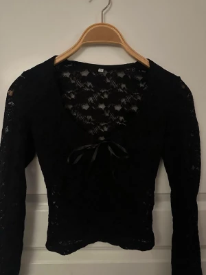 Svart spetstopp med rosett - Säljer en svart långärmad spetstopp med v-ringning och en söt rosett framtill. Toppen är genombruten med blommigt spetsmönster och har en croppad passform som ger en trendig look. Perfekt att styla med höga jeans eller kjol.