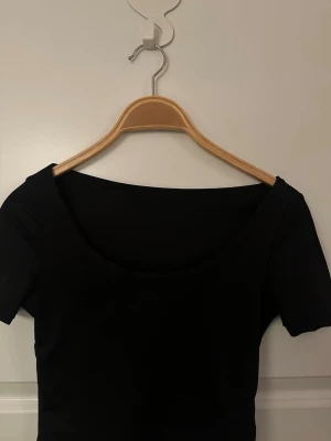 Svart basic topp med rund ringning - En svart, enkel topp med korta ärmar och rund, djup ringning. Perfekt att styla till olika outfits och har en tight passform. Tillverkad i mjukt bomullsmaterial för skön känsla hela dagen.