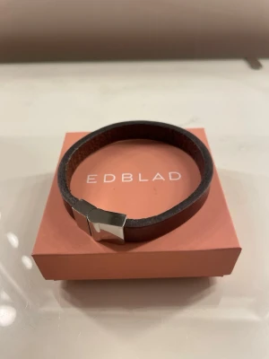 Brunt läderarmband från Edblad - Snyggt och stilrent armband från Edblad i mörkbrunt läder med ett elegant, silverfärgat metallspänne. Armbandet har en enkel och modern design som passar perfekt till en avslappnad eller trendig look. Levereras i original ask och enbart använd ett fåtal gånger. Inga defekter och nypris 500-600kr.