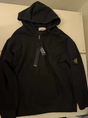 Stone Island-tröja – svart - Svart Stone Island-tröja i klassisk design med rund hals och ikonisk avtagbar badge på ärmen. Tillverkad i högkvalitativt, mjukt material som kombinerar komfort och stil. Aldrig använd – perfekt för både vardag och streetwear.