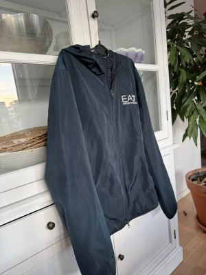 Svart vindjacka EA7 Emporio Armani - Snygg svart vindjacka från EA7 Emporio Armani med dragkedja framtill och huva. Jackan har logga tryckt på bröstet och är tillverkad i ett lätt, vindtätt material som passar perfekt för blåsiga dagar. Enkel och stilren design som funkar till det mesta.