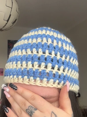 Blåvit virkad randig mössa - Säljer en handvirkad mössa med breda blå och vita ränder. Mössan har en luftig och chunky struktur som ger en cool retrovibe. Perfekt accessoar för dig som vill sticka ut och addera färg till din outfit.