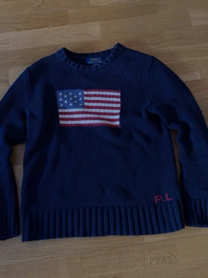 Polo Ralph Lauren Flagg Tröja - Säljer en marinblå stickad tröja från Polo Ralph Lauren med amerikanska flaggan broderad på bröstet och röda RL-detaljer vid nederkanten. Tröjan har rund halsringning och ribbstickade muddar. Perfekt för dig som gillar klassisk och preppy stil.
