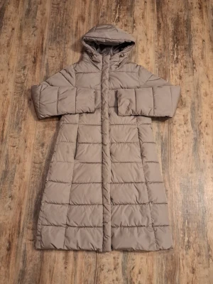 Lång beige dunjacka med huva - Snygg lång beige dunjacka med quiltat mönster och huva. Jackan har raka ärmar, dold dragkedja och tryckknappar framtill. Perfekt för kalla vinterdagar och ger en clean, stilren look.