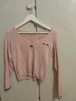 Ljusrosa kofta med broderi - Söt ljusrosa kofta från Odd Molly med v-ringning och knappar framtill. Liten mörkblå rosett vid halsen och broderad logga på bröstet. Långärmad och något croppad modell med ribbade muddar och diskret lapp nertill.