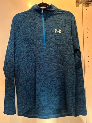 Blå långärmad träningströja Under Armour - Snygg blåmelerad långärmad träningströja från Under Armour med halv dragkedja framtill och hög krage. Tröjan har logga på bröstet och i nacken, samt är tillverkad i ett lätt och stretchigt funktionsmaterial som passar perfekt till träning.