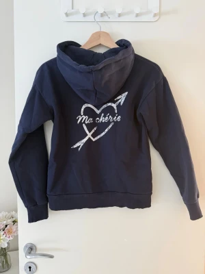 Mörkblå hoodie med tryck 'Ma chérie' - Mörkblå hoodie, På bröstet står det 'Ma chérie' i vitt och på ryggen finns ett större tryck med ett hjärta och pil samt texten 'Ma chérie'. 