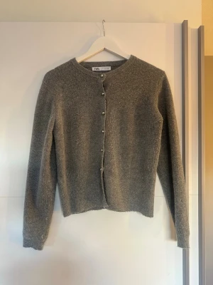 Grå kofta med pärlknappar från Zara - OBS STORLEK L MEN SITTER SOM EN S. SÅ SKULLE SÄGA ATT DET ÄR STORLEK S. Mysig grå kofta från Zara med rund halsringning och dekorativa silvriga pärlknappar framtill. Koftan har lång ärm och är stickad i ett mjukt material som känns skönt mot huden. Enkel och stilren design som passar till många olika looks.