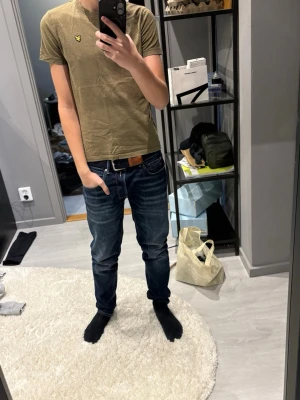 Olivgrön t-shirt från Lyle & Scott - Snygg olivgrön t-shirt från Lyle & Scott med klassisk gul logga på bröstet. T-shirten har rund halsringning och korta ärmar. Tillverkad i mjuk bomull som känns skön mot huden. Perfekt till jeans eller shorts för en avslappnad look.