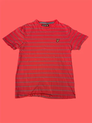 Randig röd t-shirt från Lyle & Scott - Säljer en röd t-shirt från Lyle & Scott med tunna horisontella ränder i blått och det klassiska broderade örnloggan på bröstet. T-shirten har korta ärmar och rund hals. Perfekt för dig som gillar stilrena och sportiga plagg.