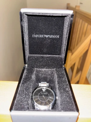 Emporio Armani silverfärgad klocka - Stilren klocka från Emporio Armani med svart urtavla och romerska siffror. Armbandet är i silverfärgad metall och klockan har en klassisk rund form. Perfekt accessoar för dig som gillar en elegant och tidlös look.