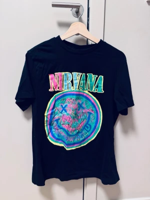 Svart Nirvana t-shirt från H&M - Cool svart t-shirt från H&M med färgglatt Nirvana-tryck framtill. T-shirten har klassisk passform och korta ärmar. Materialet är mjuk bomull som känns skönt mot huden. Perfekt för dig som gillar bandtröjor och vill sticka ut med en unik design. Ny!