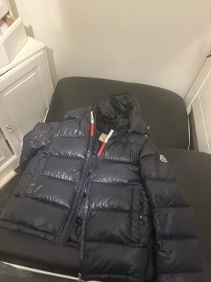 Mörkblå dunjacka från Moncler - Snygg mörkblå dunjacka från Moncler med klassisk quiltad design och stor huva. Jackan har dragkedja framtill, två stora fickor och ikonisk Moncler-logga på ärmen. Insidan har det typiska serietidningsfodret. Perfekt för kalla vinterdagar.