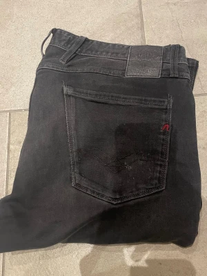 Svarta slim fit jeans från Replay - Säljer ett par svarta slim fit jeans från Replay med klassisk femficksdesign och diskret Replay-logga på bakfickan. Jeansen har en något tvättad look och smal passform som sitter snyggt längs benen. Materialet är mjukt och stretchigt för extra komfort.