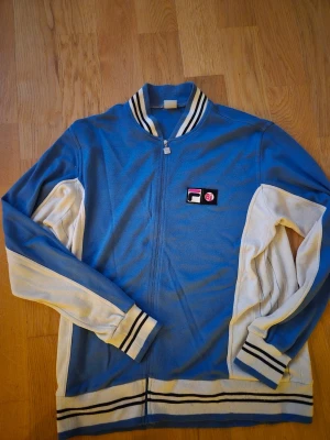 Blåvit ziptröja från Fila BJ - Snygg blå och vit ziptröja från Fila med klassisk retrostil. Tröjan har hel dragkedja, ribbade muddar och krage med svarta och vita ränder. Fila-logga på bröstet och kontrastfärgade ärmar. Perfekt för en sportig och avslappnad look. Storleken är XL, det finns tecken på användning men i sparsamt skick. Finns i Hässelby. 