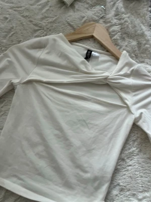 Vit topp med twistad detalj Divided - Säljer en vit långärmad topp från Divided med snygg twistad detalj framtill vid halsen. Toppen har en figurnära passform och är tillverkad i mjuk bomullsmix. Perfekt för dig som gillar stilrena plagg med en liten twist!