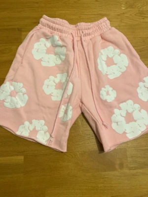 Denim tears shorts - Säljer ett par ljusrosa shorts med vita blommönster och resår i midjan. Shortsen har snörning framtill och en bakficka. Perfekta för sommaren och riktigt sköna att ha på sig. Materialet känns mjukt och bekvämt.
