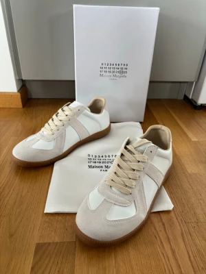 Maison Margiela Replica sneakers vita/beige - Maison Margiela Replica sneakers i vitt och beige med ovandel i läder och mocka, samt klassisk gummisula. Snygg retrostil med beige snören och diskreta detaljer. Perfekta för dig som gillar minimalistisk och stilren design.