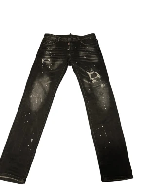 Svarta Dsquared jeans med färgstänk - Säljer ett par svarta jeans med coola slitningar och vita färgstänk för en edgy look. Jeansen har rak passform och klassisk femficksdesign. Perfekta för dig som gillar streetwear och vill sticka ut lite extra.