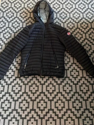 Svart dunjacka med huva från Colmar - Snygg svart dunjacka från Colmar med huva och två dragkedjeförsedda fickor. Jackan har ett quiltat mönster och den klassiska Colmar-loggan på bröstet. Perfekt för kalla vinterdagar och riktigt skön att bära.