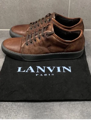 Bruna sneakers från Lanvin - Snygga bruna sneakers från Lanvin i läder med svarta detaljer och svart sula. Klassisk låg modell med rund tå och bruna skosnören. Perfekta för dig som gillar stilrena och exklusiva sneakers med en tidlös vibe.