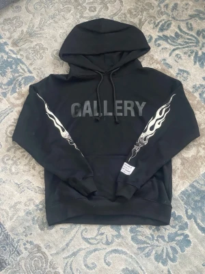 Svart Gallery Dept hoodie med flames - [FRI FRAKT 🚚] Snygg svart hoodie från Gallery Dept med stora flames längs båda ärmarna och trycket 'GALLERY' på bröstet. Klassisk känguruficka, dragsko i huvan och ribbade muddar. Perfekt för dig som gillar streetwear och vill sticka ut lite extra.