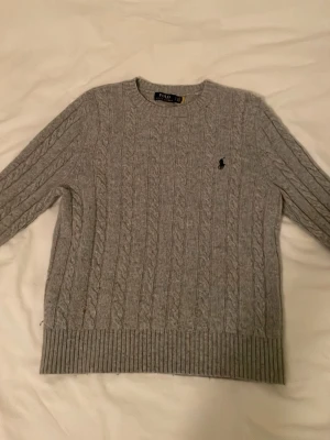 Grå kabelstickad tröja Polo Ralph Lauren - Kabelstickad grå tröja från Polo Ralph Lauren med klassisk svart broderad logga på bröstet. Tröjan har rund halsringning, ribbade muddar och är perfekt för dig som gillar en tidlös och clean stil.