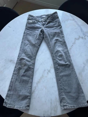 Grå bootcut jeans med fickdetaljer - Säljer ett par grå bootcut jeans med snygga fickdetaljer och knappar bak. Jeansen har låg midja och klassisk femficksdesign. 