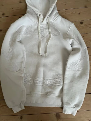 Vit hoodie från Burberry med brodyr - Säljer en clean vit hoodie från Burberry med snygg broderad logga och bladmönster på bröstet och ärmarna. Hoodien har dragsko i huvan och ribbade muddar. Perfekt för dig som gillar exklusiv streetwear och vill sticka ut med stilren design. Lite stor i storleken