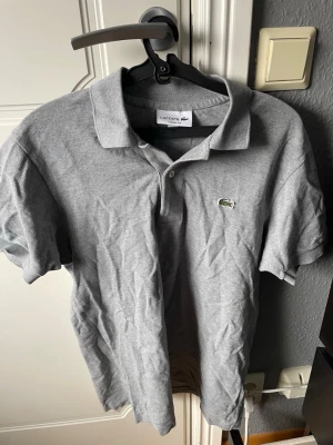 Grå pikétröja från Lacoste - Klassisk grå pikétröja från Lacoste med den ikoniska gröna krokodilloggan på bröstet. Tröjan har korta ärmar, krage och knappar vid halsen. Tillverkad i mjuk bomull och har en normal passform. Perfekt för en clean och stilren look.